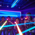 Children and adults playing laser tag in an illuminated indoor arena with colorful laser beams.